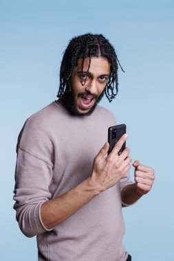 Heyecanlı Arap adam akıllı telefondan mesajları kontrol ediyor ve kazanan hareketi yumruğunu sıkarak gösteriyor. Cep telefonu kullanan ve mutlu bir ifadeyle kameraya bakan kişi