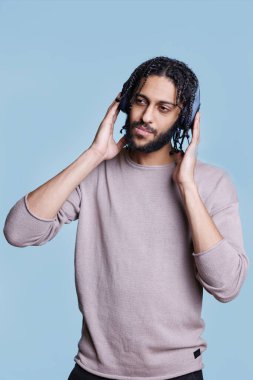 Arap adam şarkı dinliyor, kablosuz kulaklıklara dokunuyor ve dalgın bir yüz ifadesiyle yüzünü çeviriyor. Kulaklık takan, günlük kıyafetler giyen, müzikten hoşlanan biri.