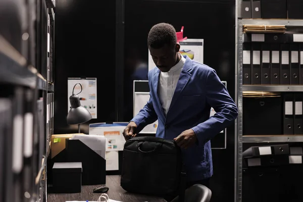 African american detective preparing his backpack, leaving job late at night in arhive room. Police officer working overtime at criminal case, analyzing crime scene evidence. Criminology concept