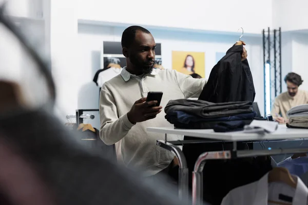 アフリカ系アメリカ人男性が衣料品店でカジュアルなジャケットを選び 携帯電話を使ってウェブサイトで価格を確認しています 百貨店のクライアントがアパレルラックを閲覧し ブティックアプリを調べる — ストック写真