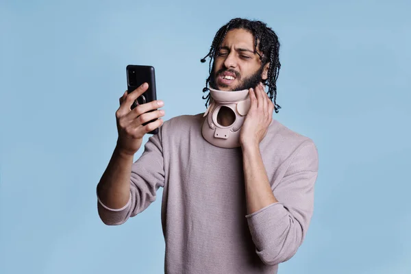 Arab man receiving medical advice through video call on mobile phone while holding neck brace. Young person consulting with doctor online on smartphone about injury recovery