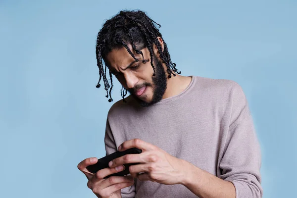 重点是拿着手机玩网络游戏的年轻Arab男子 面部表情紧张的人喜欢智能手机上的娱乐性应用软件 — 图库照片