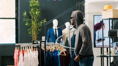 Afro-Amerikan hırsızı elbise mağazasında güvenlik görevlisi tarafından yakalanarak moda kıyafetleri çalmaya çalışıyor. Asyalı koruma soyguncunun modaya uygun malları iade etmesini yoksa polisi arayacağını söyledi.