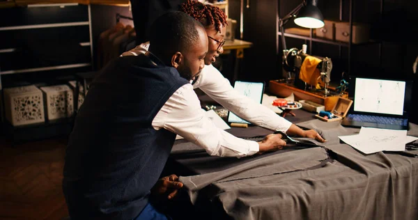 African american tailors cutting garment, working in team to create custom made tailored clothes. Craftsman and assistant using tailoring tools in atelier, preparing for needlework. Handheld shot.
