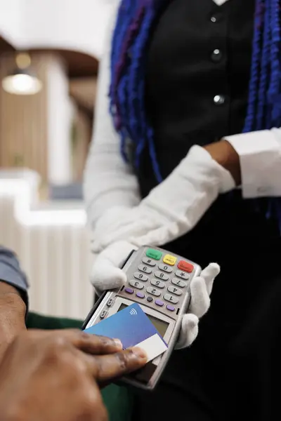 Man making payment using contactless credit card, closeup of hand paying bill order with NFC technology while resting at hotel lounge area. Waitress wearing white gloves holding POS terminal