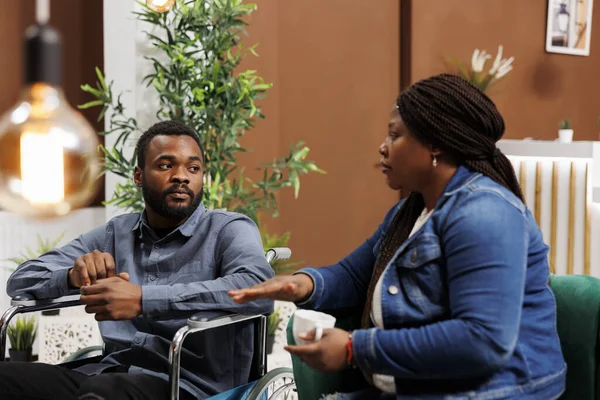African American couple arriving at rehabilitation center for disabled, young man wheelchair user traveling with wife, sitting together in hotel lobby waiting for room with disability access
