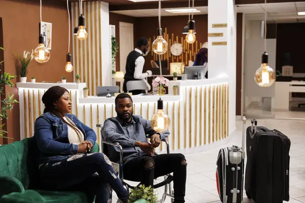 年轻的非洲裔美国残疾人轮椅使用者与私人助理 残疾人和他的妻子坐在酒店大堂与行李同行 为残疾人提供的客人体验 — 图库照片
