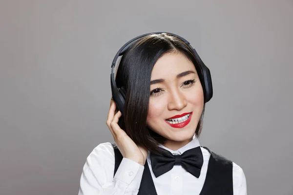 穿着制服的亚洲女服务员在无线耳机里听音乐 有魅力的餐厅接待员 在演播室里摆姿势时 戴着耳机 欣赏着播放列表 — 图库照片
