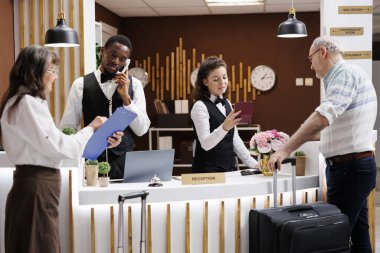 Resepsiyondaki yaşlı erkek turistin kimliğini kontrol ederken Afro-Amerikan personeli telefona cevap veriyor. Emekli kadın evrakları doldururken kıdemli adam pasaportunu bekliyor..