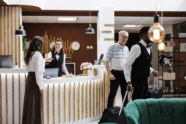 stock image Female concierge assists elderly lady at front desk, providing smooth check-in process and verifying reservations. African american bellboy helps retired senior man with suitcases to hotel room.