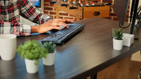 Employee remotely working from home on computer, inputing data in his brick wall apartment. African american freelancer typing on keyboard, teleworking at nice clean wooden desk, close up