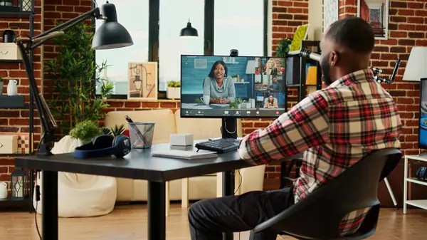 Smiling employee in video conference meeting greeting remote working coworkers. Man teleworking from his stylish apartment in online video call with diverse international team