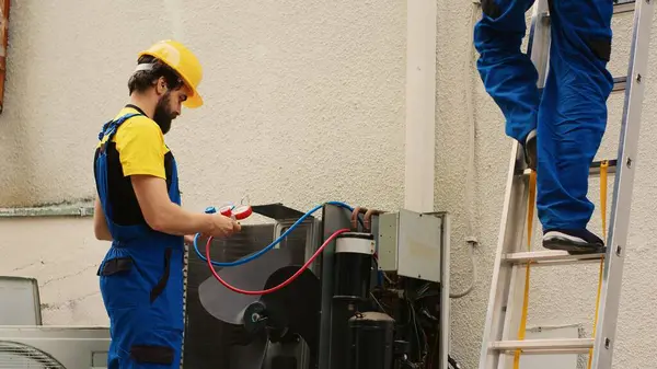 Expert mechanic measuring refrigerant levels in air conditioner with manifold gauges while african american coworker steps down from folding ladder after finishing checking rooftop HVAC system