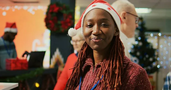 Portrait of smiling employee wearing Santa hat walking through clothing rack rows in xmas adorn shopping mall store, using clipboard to crosscheck price tags, making sure shop is ready for customers