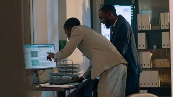 African american businessmen trying to troubleshoot reason for company revenue down from last year, checking numbers in office. Employees looking over financial charts during nightshift