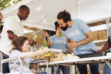 Dost canlısı erkek gönüllü, tekerlekli sandalyedeki yoksul kadına yardım ediyor ve bedava yemek veriyor. Bir açık hava gıda bankasında hayır işçileri engellilere ve daha az talihlilere açlık yardımı yapıyor..