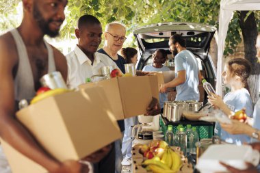 Yemek kutuları ve konserve yiyecekleri ihtiyacı olanlara dağıtırken gülümseyen bir grup birey. Genç gönüllüler, barınak arayan yaşlılara ve bireylere memnuniyetle insani yardım sağlıyorlar..