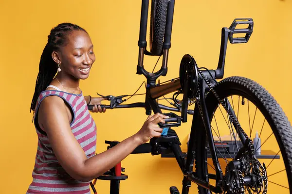 Active young female cyclist ensures bike is well-prepared for outdoor adventures by diligently examining the pedals and gear. Sporty african american lady grasping bicycle foot lever for maintainance.