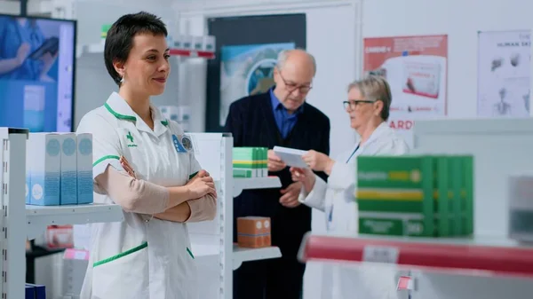 stock image Friendly pharmacist in apothecary placing pills box on shelves while senior colleague helps customer with informations. Smiling wellness expert in drugstore arranging medicinal items