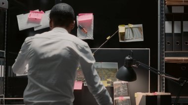 Male inspector analyzing evidence on board in archive room, reading witness statements. Private office looking at suspicious photos and checking criminal records in investigation office.