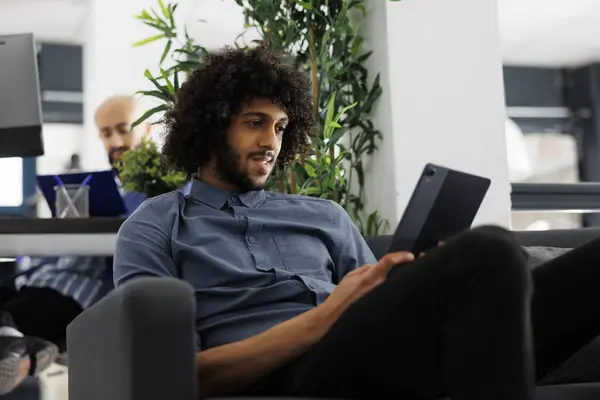 Arab entrepreneur checking financial report on digital tablet while working in business office. Start up company worker browsing internet while sitting on couch in coworking space