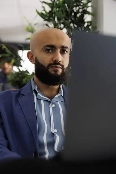 Arab businessman sitting in front of laptop at desk, preparing for video call with clients. Executive manager checking financial analytics report before presentation in coworking space