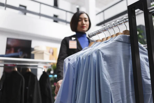 蓝色的休闲衫挂在衣架上 而亚洲的女店员则站在购物中心的服装店里选择性地聚焦 省中心精品店特写镜头衣架上的时髦服装 — 图库照片