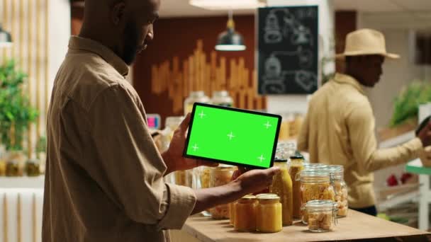 アフリカ系アメリカ人のバイヤーは 地元の食料品店で緑色のテンプレートが付いているタブレットを示し 化学物質のフリーアイテムの隣の空白のコピースペースを見ます 通常のクライアントは 染色体を持つデバイスを保持しています カメラ — ストック動画