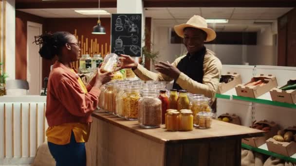 农民与围裙推荐新的自制酱汁的妇女 显示有机面食类型和其他食品供应 非洲裔美国人卖主向常客介绍产品 摄像头B — 图库视频影像