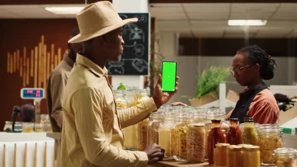 アフリカ系アメリカ人セラーがグリーンスクリーンでスマートフォンアプリを保持し 地元のスーパーマーケットで隔離されたモックアップテンプレートを提示しています ビーガン商人は有機食料品店で染色レイアウトを使用しています カメラ — ストック動画