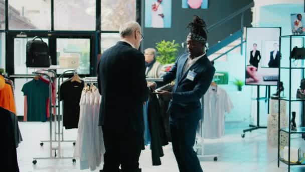 アフリカ系アメリカ人の店のアシスタントは ショッピングセンターのスーツで定期的なクライアントを助け 正式な服を購入しようとシニアの顧客 高齢者はショッピングモールで新しいコレクションを買う カメラ — ストック動画