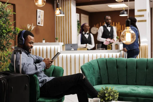 快乐微笑的黑人小伙子坐在酒店大堂的无线耳机里 用数字平板电脑 年轻的非洲裔美国人在等待入住时正在看电影 — 图库照片