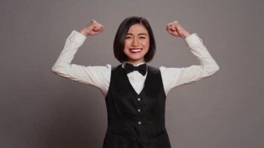 Asyalı otel kapıcısı gri stüdyoda kol kaslarını esnetiyor, kamera önünde gücünü ve gücünü gösteriyor. Fiyonklu ve üniformalı bir resepsiyonist kasları esnek duruyor. Kamera B.