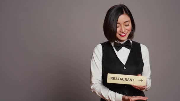 アジアのホテルのコンシェルジュは ダイニングエリアを指す方向を示すレストランのサインを保持しています すべてのアメニティを楽しむためにクライアントを支援するレセプションは 灰色の背景の上に立っています カメラ — ストック動画
