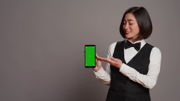 アジアのホテルのコンシェルジュは 緑色のレイアウトで電話を指し 隔離された染色体テンプレートを実行しているデバイスを保持しています コピースペースモックアップでスマートフォンディスプレイを表示するレセプション カメラ — ストック動画