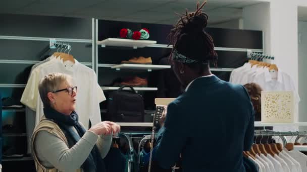 精品店的助理帮助老年妇女选择名牌包包 在时装展示室里查看多个品牌和模特 非洲裔美国工人向客户展示时髦的配件 相机A — 图库视频影像