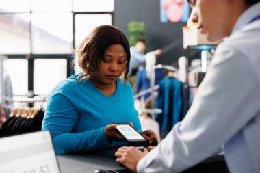 Moda mağazası masasında şık kıyafetler alan şık bir kadın, NFC ve kredi kartı olan bir cep telefonu. Modern butikte resmi kıyafetler için müşteri alışverişi.