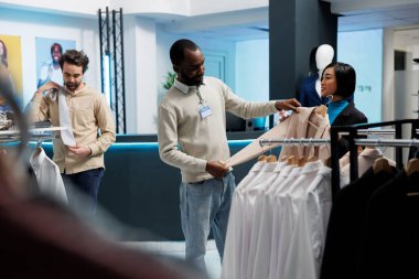 Gülümseyen Asyalı kadın ceketini tutuyor ve kıyafet alışverişi yaparken butik danışmanından moda tavsiyesi istiyor. Giyim mağazası müşterisi ve işçi kadın giyim kuşamını tartışıyor