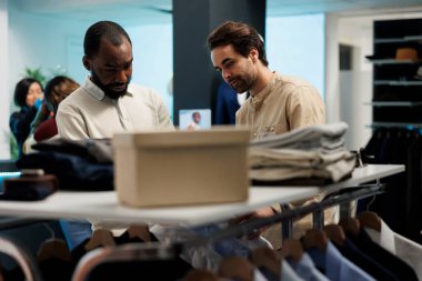 Müşteri ve asistan elbise raflarını incelerken kıyafetleri tartışıyor. Alışveriş merkezi butik alışverişçisi satın almadan önce giysileri incelerken çalışandan rehberlik alıyor