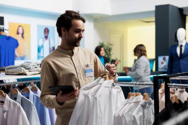 Gülümseyen beyaz bir adam giyim mağazasında çalışırken ürünlerini seriyor. Alışveriş merkezi butik çalışanı dijital tablet tutuyor ve raflarda moda kıyafetler asıyor.