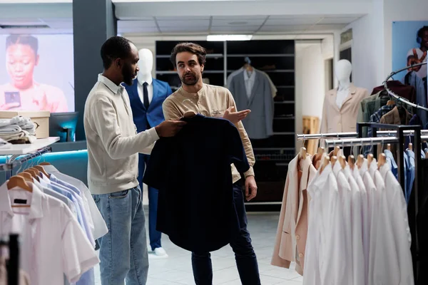 Askıda tişört tutan Afrikalı Amerikalı bir adam alışveriş merkezi çalışanına boyut seçeneklerini soruyor. Butik işçisi, yeni koleksiyon rafını göstererek müşteriye kıyafet seçmesinde yardımcı oluyor.