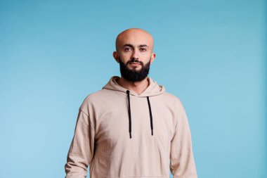 Genç Arap adam kameraya bakarken rahat poz veriyor. Tarafsız yüz ifadesine sahip kel sakallı adam. Mavi arka planda bej kapüşonlu kıyafet stüdyo resmi giyiyor.