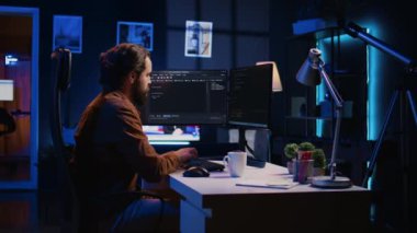 Neon lit ev ofisinde uzaktan iş değişimi sırasında web geliştirme programcısı, kodlama projesini bitiriyor. Bilgisayar masasında oturan bilişim uzmanı, masaüstü bilgisayarı, kamera B üzerinde çalışıyor.