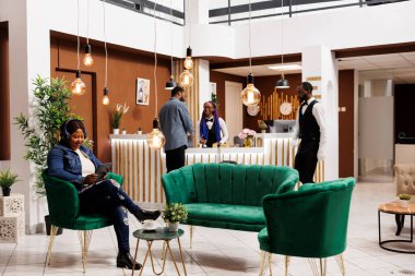 Afro-Amerikan kadın otel misafiri, kulaklık takmış dijital tablete bakıyor lobide dinleniyor. Sırıtkan siyah kadın gezgin kayıt için beklerken internetten film izliyor.