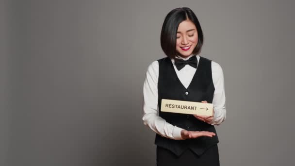 アジアのホテルのコンシェルジュは ダイニングエリアを指す方向を示すレストランのサインを保持しています すべてのアメニティを楽しむためにクライアントを支援するレセプションは 灰色の背景の上に立っています カメラ — ストック動画