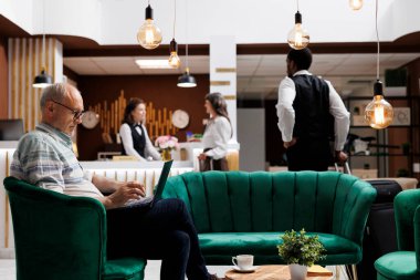 Emekli son sınıf öğrencisi, otel lobisinden tatilini planlarken online tatil aktivitelerine erişmek için bilgisayarını kullanıyor. Profesyonel çalışanlar resepsiyonda yaşlı kadın yolculara yardım ediyor..