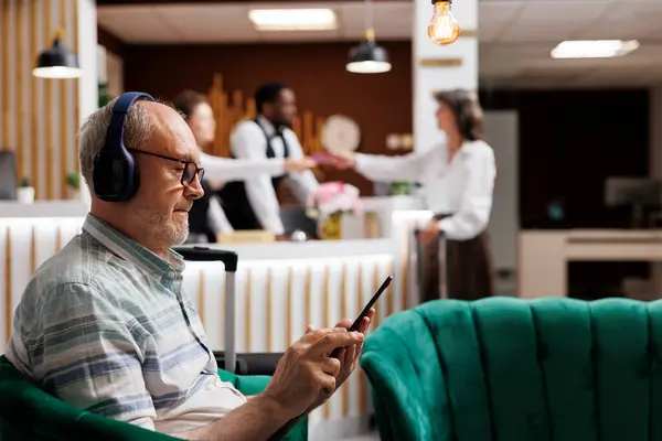 Detailed view of elderly male guest relaxing in cozy lounge area with tablet and wireless headset. Retired old man listening to music and surfing the net on digital device. Close-up, side-view.