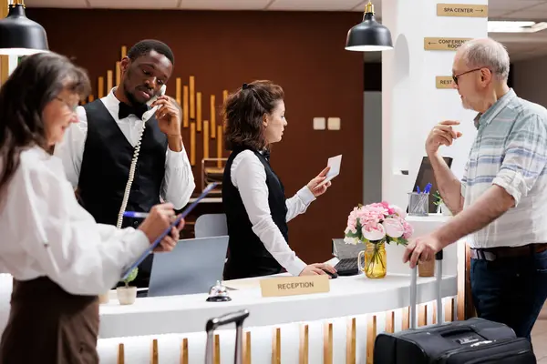 While african american staff answers phone, female concierge verifies id of elderly male traveler at hotel front desk. Retired senior woman fills out form as old man waits for his passport at counter.