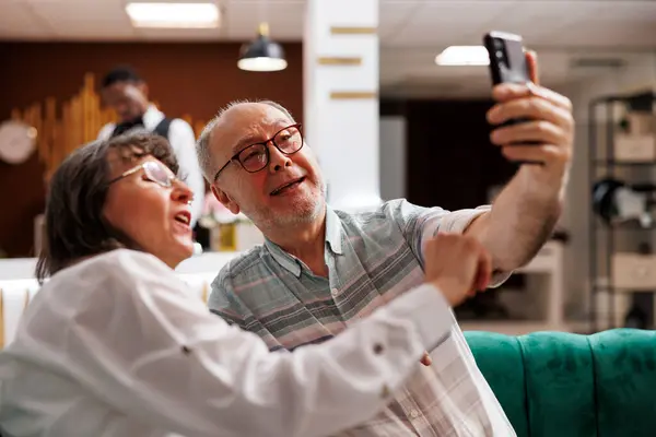 高齢のカップルは ビデオ通話のためのモバイルデバイスを使用して 豪華なホテルのロビーで休暇を楽しんでいます ラウンジエリアに座り 家族とスマートフォンで話す上級旅行者 — ストック写真