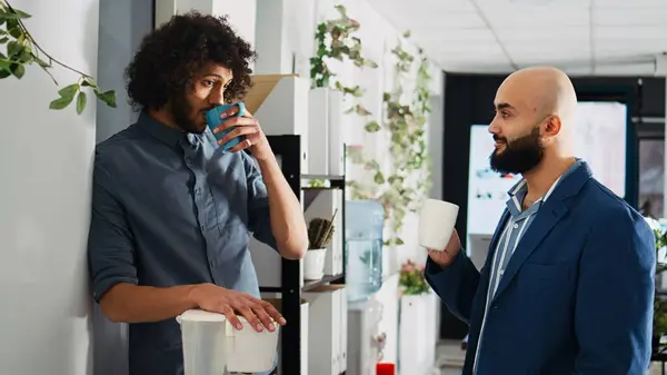Employees relaxing on coffee break, laughing together and having fun before starting to work on small business. Two partners talking to each other, standing in coworking space.